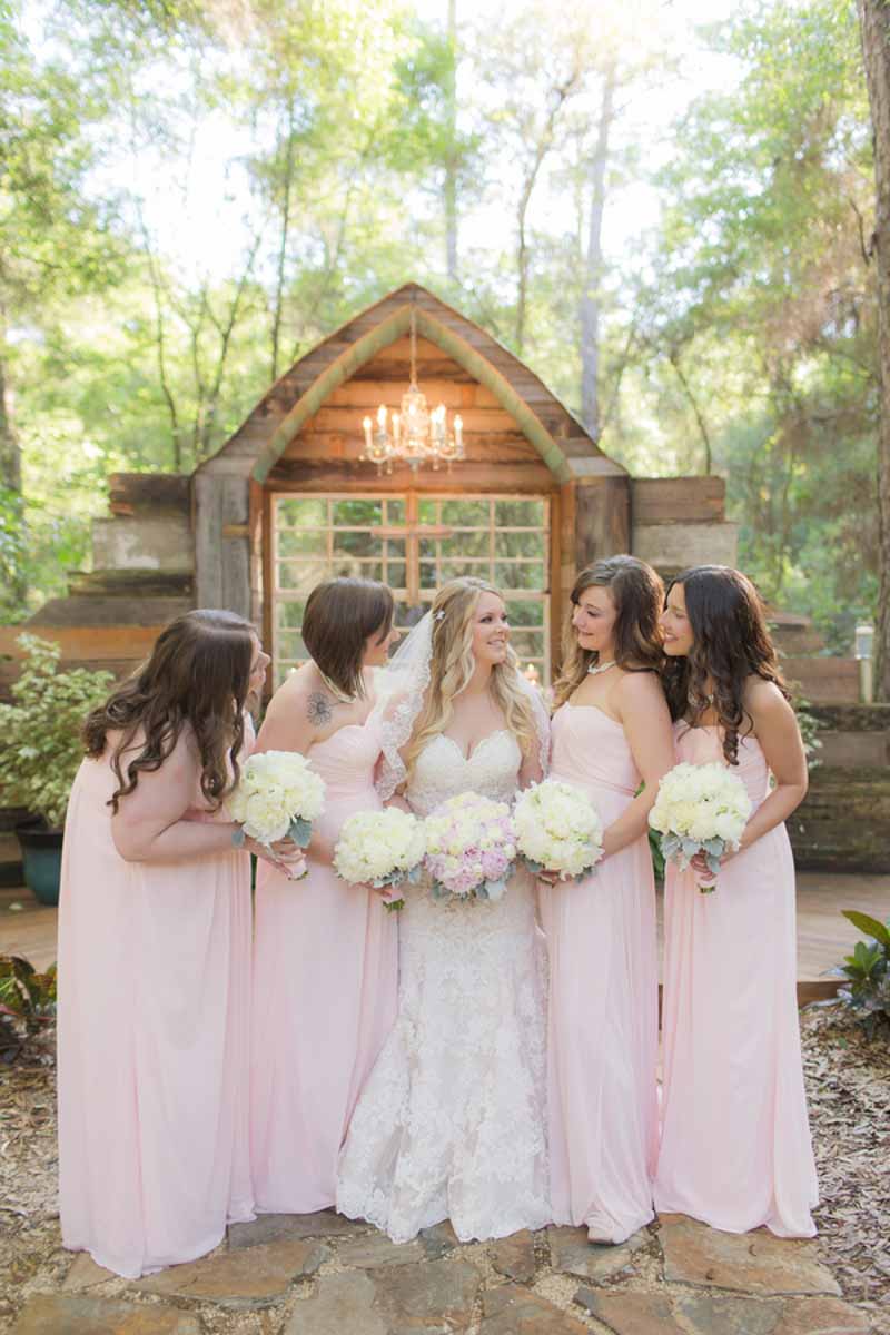 bridesmaids in blush gowns looking at bride holding bouquets in front of Bridle Oaks wedding ceremony space