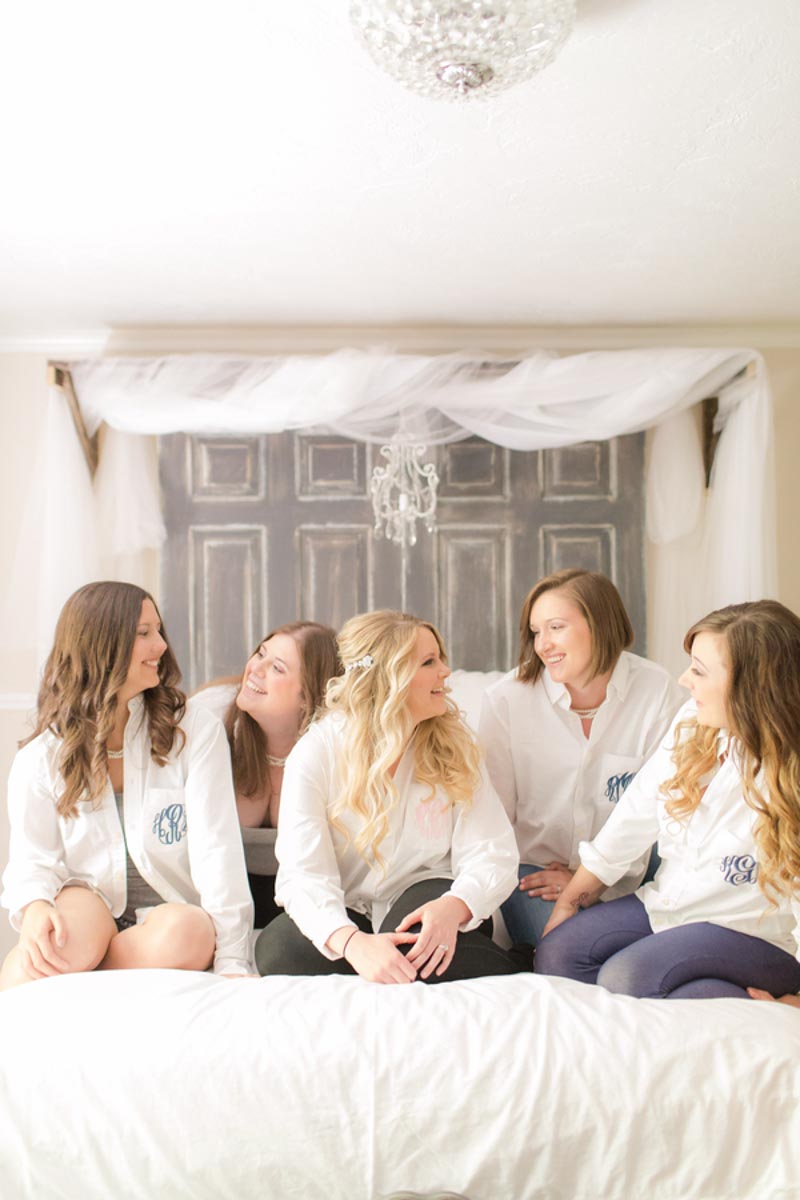 bride and bridesmaids on bed smiling at each other in monograms shirts