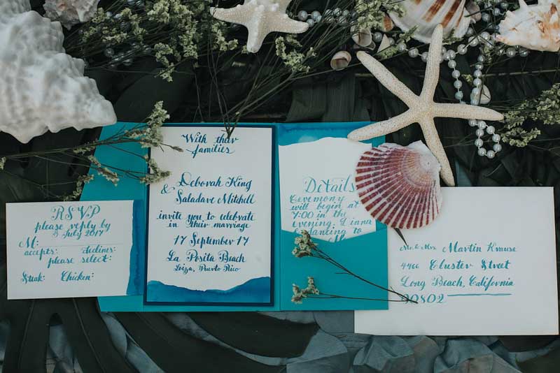 blue and teal calligraphy wedding invitations surrounded by seashells