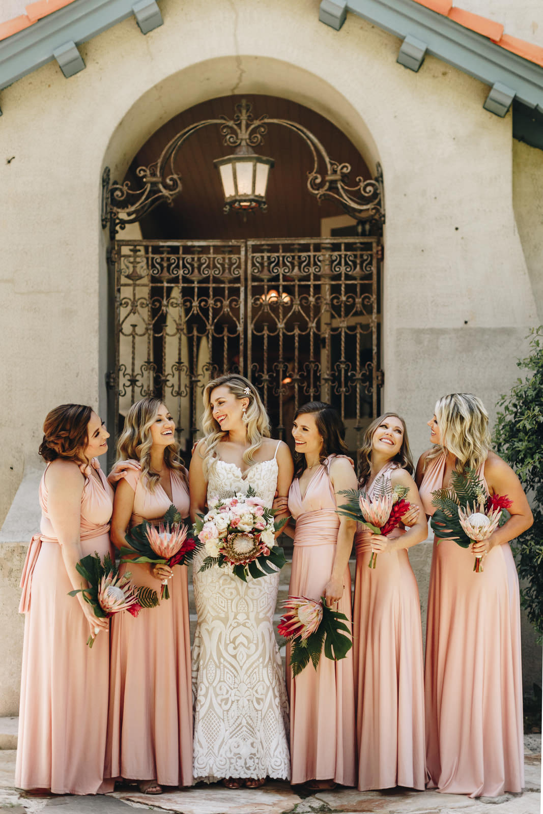 Bridesmaids with the bride, looking at each other and holding bouquets with a king protea. Bridesmaids are wearing blush dresses. 